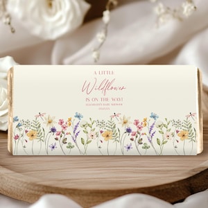 May include: A rectangular baby shower favor with a cream background and floral border. The text "A Little Wildflower is on the way!" is written in a script font. The floral border features colorful wildflowers and dragonflies.