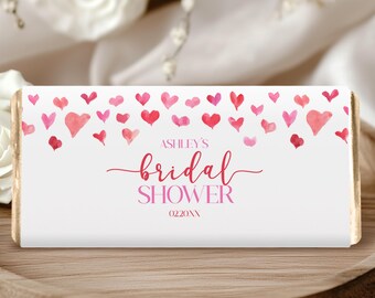 May include: A white candy bar wrapper with a pink heart design and the words "Ashley's Bridal Shower" in pink script. The date "02.20XX" is below the text. The candy bar is on a wooden tray with white roses in the background.