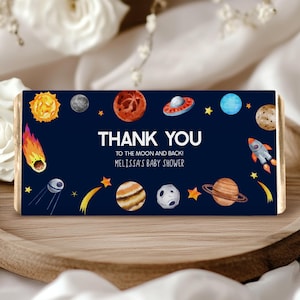 Puede incluir: Un envoltorio de chocolate de agradecimiento azul y amarillo con temática espacial. El envoltorio presenta planetas, estrellas, una nave espacial y el texto "¡Gracias hasta la luna y de regreso! Baby shower de Melissa".