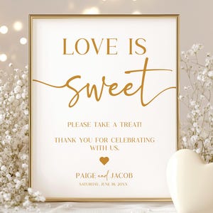May include: A framed sign with the text "LOVE IS sweet" in gold script. The sign also includes the text "PLEASE TAKE A TREAT! THANK YOU FOR CELEBRATING WITH US." A white heart-shaped object is on the right.