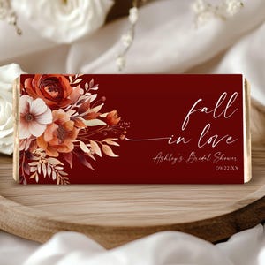May include: A rectangular chocolate bar wrapper in deep burgundy featuring a floral arrangement of rust, orange, and cream-colored flowers on the left. The text reads fall in love, Ashley's Bridal Shower, and the date 09.22.XX in white script.