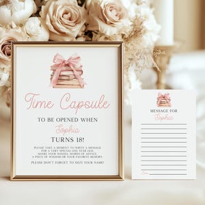 以下が含まれることがあります： 「Time Capsule」と「Sophia Turns 18!」というピンク色の筆記体で書かれた額入りのサイン。メッセージの書き方の指示と、ピンクのリボンで結ばれた本の水彩イラストが含まれています。お揃いのメッセージカードが隣にあります。