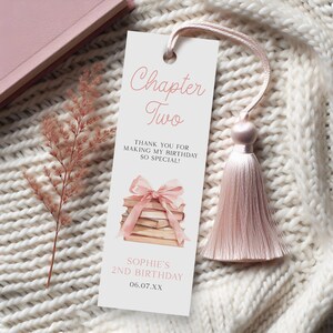May include: A rectangular bookmark with a pink tassel. The bookmark features the text "Chapter Two" and "Thank you for making my birthday so special!" A watercolor illustration of books with a pink bow is also present.