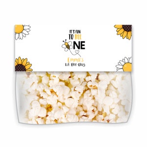 Puede incluir: Una bolsa de regalo blanca con un diseño de abeja amarilla y negra y el texto "It's fun to bee one" y "Emma's 1st Bee-Day". La bolsa está llena de palomitas de maíz.