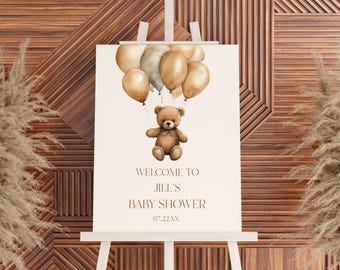 May include: A baby shower sign on a white easel. The sign features a watercolor illustration of a teddy bear holding onto balloons. Text on the sign reads "WELCOME TO JILL'S BABY SHOWER 07.22.XX". The background is a wooden wall.