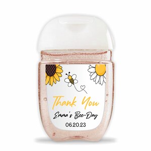 May include: A small clear bottle of hand sanitizer with a white label. The label has a sunflower, a bee, and the text "Thank You Emma's Bee-Day 06.20.23".
