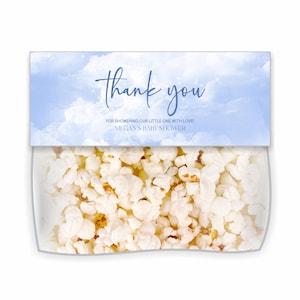 May include: A clear plastic container filled with popcorn, topped with a blue and white card. The card reads "thank you" in cursive, with additional text below. The background of the card has a cloudy sky design. The text on the card says "FOR SHOWERING OUR LITTLE ONE WITH LOVE! MEGAN'S BABY SHOWER."