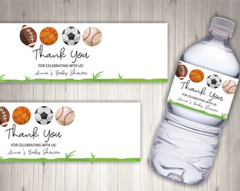 May include: Water bottle label with a green grass background and four sports balls: a football, basketball, soccer ball, and baseball. The label says "Thank You for celebrating with us! Anna's Baby Shower".