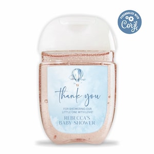 Puede incluir: Una botella de gel hidroalcohólico azul y blanca con una etiqueta de agradecimiento. La etiqueta presenta un globo aerostático y el texto "¡Gracias por duchar a nuestro pequeño con amor! Baby Shower de Rebecca".