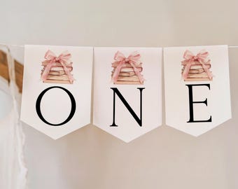 May include: A decorative banner spelling "ONE" across three white pennants. Each pennant displays a watercolour illustration of a stack of books adorned with a pink bow. The letters are black.