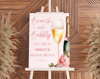 May include: A pink and white welcome sign for a birthday brunch with the text "Brunch and Bubbly" and "Welcome to Ashley's Birthday Brunch 09.20.XX". The sign features a watercolor illustration of a champagne flute and a bottle of champagne with a pink rose.