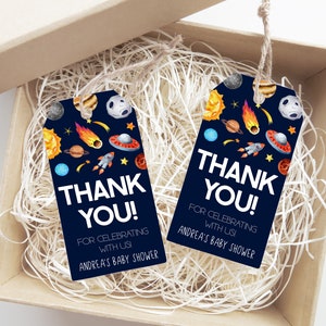 May include: Two navy blue gift tags with a space theme, featuring planets, rockets, and the text "THANK YOU!" for a baby shower. The tags are inside a cardboard box with shredded paper.
