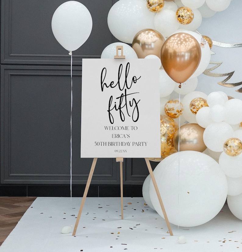 May include: White welcome sign with black calligraphy that reads "hello fifty" and the text "Welcome to Erica's 50th Birthday Party 09.22.XX". The sign is on a wooden easel in front of a wall of white and gold balloons.