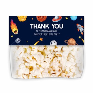May include: A blue and black thank you bag filled with popcorn. The bag has a label with a space theme and the text "Thank You To the Moon and Back! Jackson's Birthday Party".