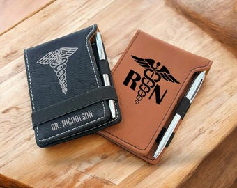 May include: Two leatherette notebooks, one black and one brown, with silver pens. The black notebook has a caduceus symbol and the name "DR. NICHOLSON". The brown notebook features the letters "RN" and a caduceus symbol.