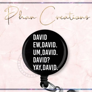 May include: A black retractable badge reel with the text "DAVID EW, DAVID. UM, DAVID. DAVID? YAY, DAVID." in white. The reel has a silver clip and a black cord. The background is a pink marble design with gold text that says "Phan Creations".