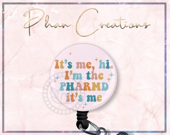 May include: A pink retractable badge holder with a white circle featuring the text "It's me, hi. I'm the PHARM'D it's me" in colorful retro font with gold glitter accents.