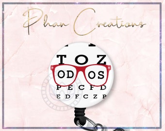 May include: White round badge with red glasses and the text "TOZ OD OS PECFD EDFCZP". The badge is attached to a black retractable badge reel.