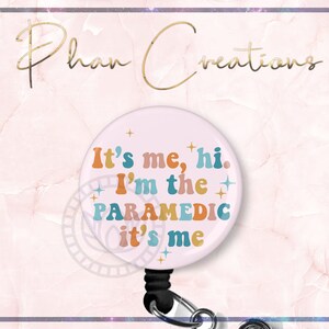 May include: A light pink badge reel with the text "It's me, hi. I'm the PARAMEDIC it's me" in retro colors. The badge reel has a black retractable cord and a silver clip. The background is a light pink marble design.