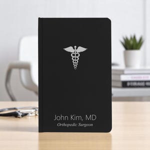 May include: Black notebook with a silver medical symbol and the text "John Kim, MD Orthopedic Surgeon".