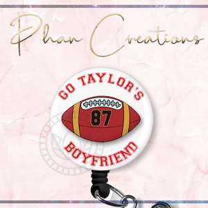 May include: White retractable badge holder with a round button featuring a red and yellow football with the text "GO TAYLOR'S BOYFRIEND" and the number "87" on it.