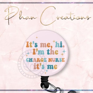 May include: A pink retractable badge holder with a white circle featuring the text "It's me, hi. I'm the charge nurse it's me" in colorful retro font with gold stars.