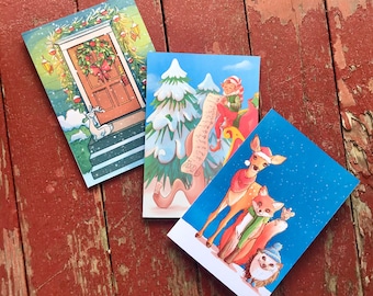 以下が含まれることがあります： イラストが描かれた3枚のクリスマスカード。1枚はトナカイが描かれた装飾されたドア、もう1枚はリストを持ったエルフ、3枚目は雪景色の中の鹿、キツネ、ハリネズミが描かれています。カードは風化した赤い木の表面に置かれています。