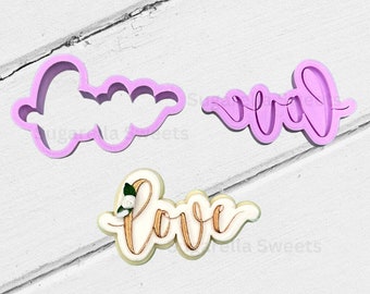 May include: Two purple plastic cookie cutters in the shape of the word "love" and a white cookie decorated with gold icing and the word "love" with a small white flower design.