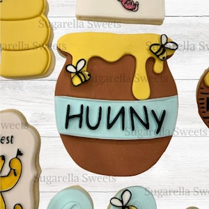 May include: A brown cookie shaped like a honey pot with yellow honey dripping from the top. The word "HUUNY" is written in black on a light blue band. Two yellow and black bees are on the cookie. Other cookies are in the background.