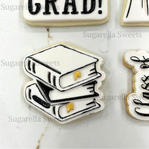 May include: A white cookie decorated with a black outline of a stack of three books. The books have gold accents on the spines.
