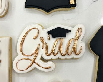 May include: A white cookie decorated with gold icing and the word "Grad" with a small black graduation cap on top.