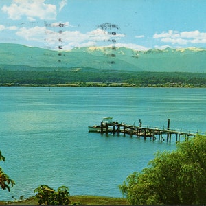 Peut inclure: Une jetée en bois s'étend sur un lac bleu calme avec un petit bateau amarré. Des arbres verdoyants bordent le rivage et des montagnes enneigées se dressent au loin.