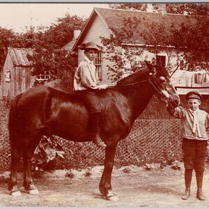 Peut inclure: Photographie vintage sépia de deux garçons et un cheval. Un garçon est assis sur le cheval, portant un chapeau et une chemise claire. L'autre garçon se tient à côté du cheval, portant une casquette et un pantalon sombre. Une maison et une clôture sont en arrière-plan.