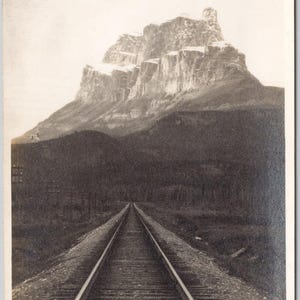 May include: Vintage black and white postcard featuring a mountain landscape with a railway track leading towards the mountain. The mountain is snow-capped. Text at the bottom reads "64. CASTLE MTN. BANFF."