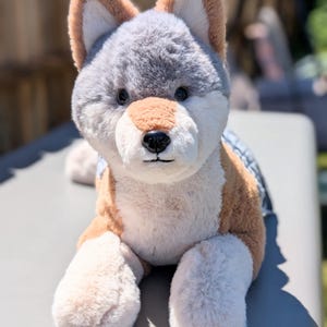 May include: A plush stuffed animal resembling a wolf or husky dog with soft fur in shades of light brown, white, and grey. The toy features pointed ears, dark plastic eyes, a black nose, and a small stitched mouth, with a grey patterned fabric visible.