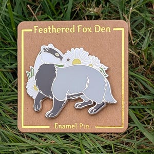 Könnte beinhalten: Ein Emaille-Pin mit einem Dachs in Grau- und Schwarztönen, verziert mit weißen Gänseblümchen mit gelben Zentren. Der Pin befindet sich auf einer braunen Karte mit dem goldenen Text "Feathered Fox Den" und "Enamel Pin".