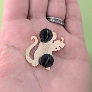 Puede incluir: Pin de metal dorado de un mono de dibujos animados sosteniendo un racimo de uvas verdes. El pin tiene dos cierres de embrague de goma negra.