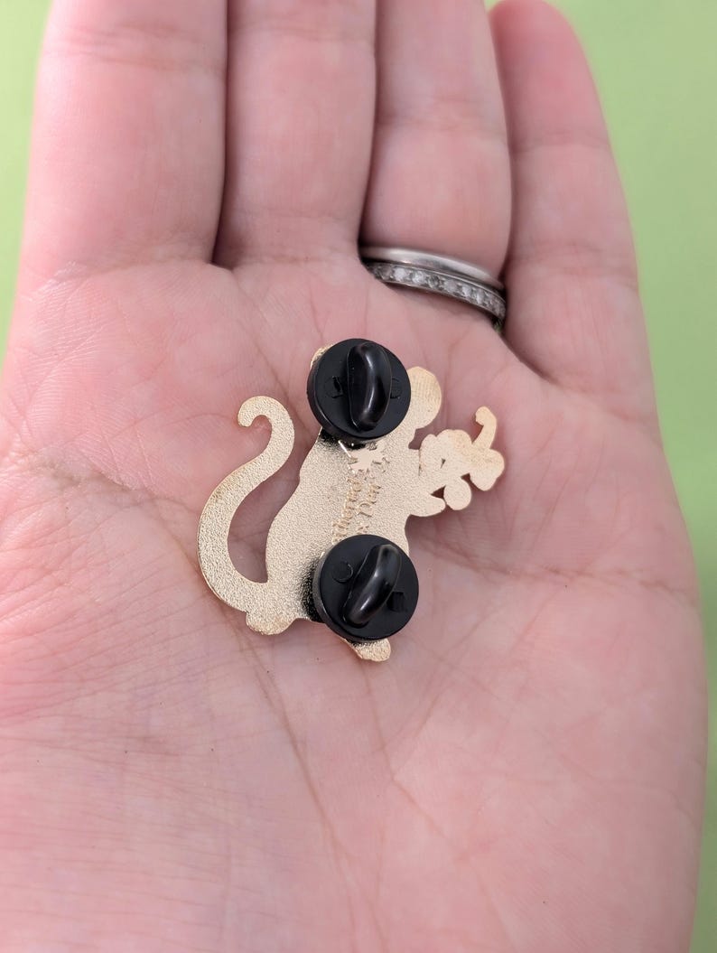 Puede incluir: Pin de metal dorado de un mono de dibujos animados sosteniendo un racimo de uvas verdes. El pin tiene dos cierres de embrague de goma negra.
