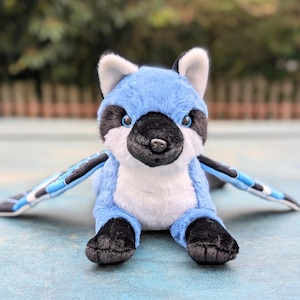 May include: A plush blue jay stuffed animal with outstretched wings. The toy has a black and white face, black feet, and blue wings with black and white accents. The plush toy is designed to look like a bird.