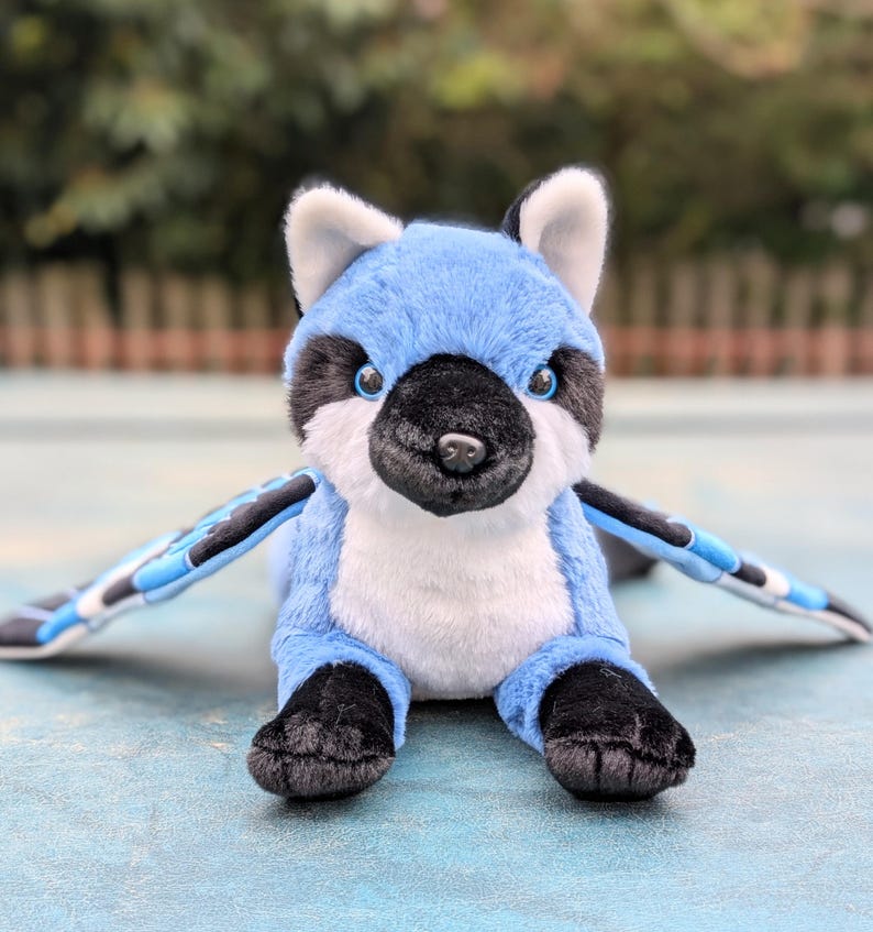 May include: A plush blue jay stuffed animal with outstretched wings. The toy has a black and white face, black feet, and blue wings with black and white accents. The plush toy is designed to look like a bird.