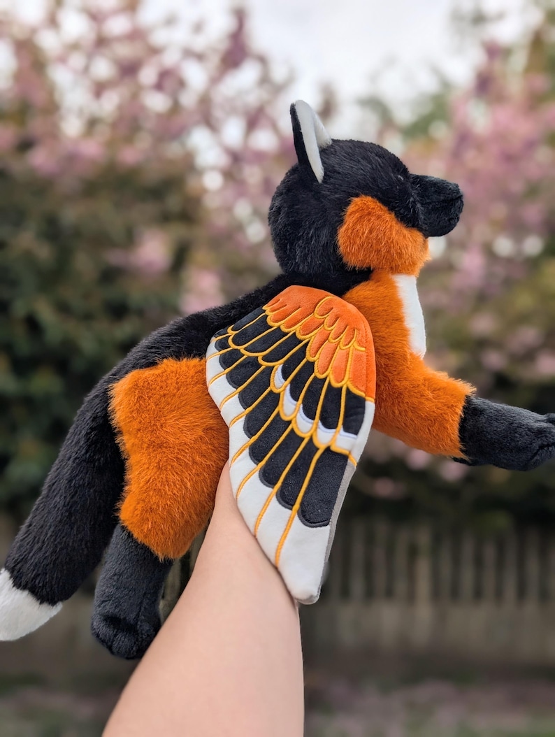 May include: A plush fox toy with black, orange, and white fur, and large, detailed wings. The wings feature a black and white pattern with orange accents. The fox is held up against a blurred background of pink blossoms.