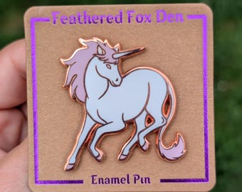 May include: A rose gold enamel pin featuring a white unicorn with a pink mane and tail. The pin is on a brown card with the text "-Feathered Fox Den-" and "Enamel Pin" in purple.