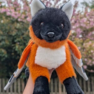 May include: A plush toy fox with black fur on its head and paws, and orange fur on its body. The fox has white accents on its chest and ears, and gray and orange wings. The toy is designed to look like a fox with wings.