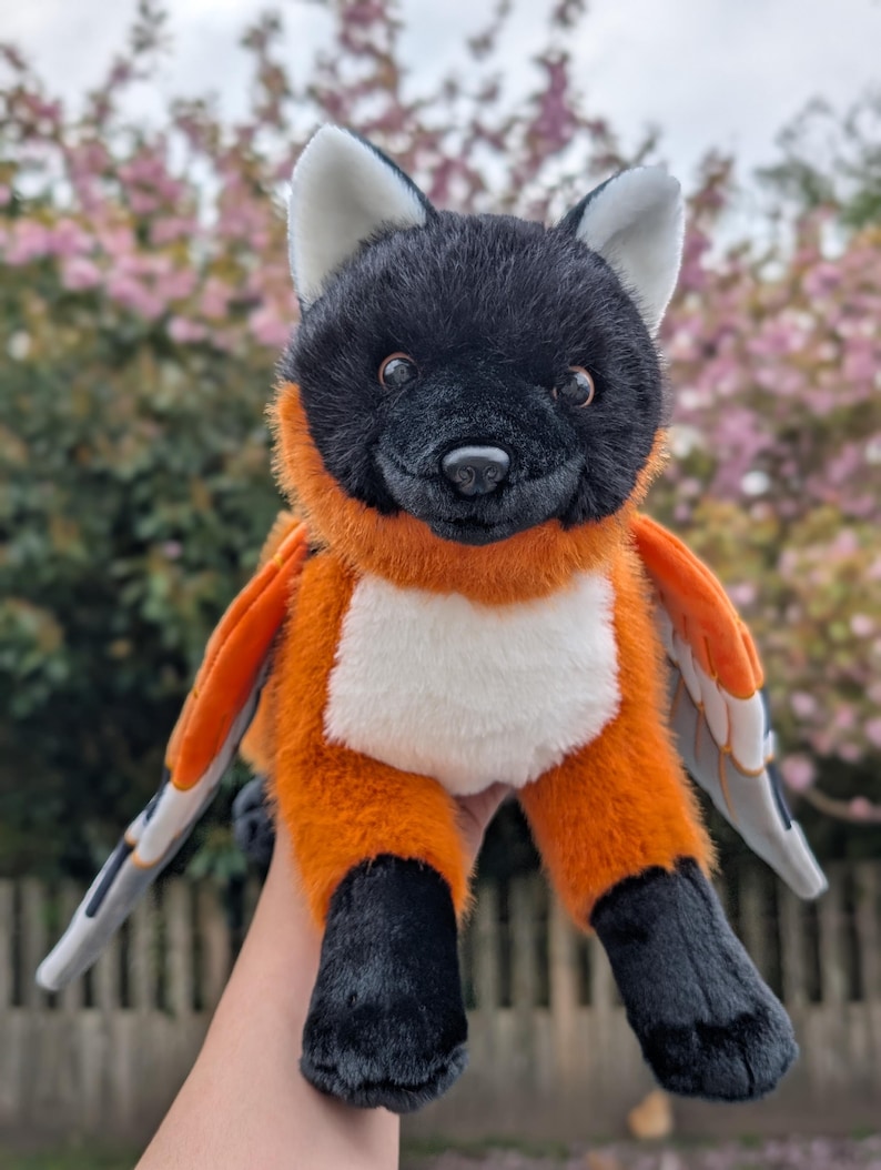 May include: A plush toy fox with black fur on its head and paws, and orange fur on its body. The fox has white accents on its chest and ears, and gray and orange wings. The toy is designed to look like a fox with wings.