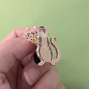 Puede incluir: Pin de esmalte dorado con un rat&oacute;n de dibujos animados sosteniendo una rama de bayas moradas. El rat&oacute;n tiene la nariz y la cola rosas y est&aacute; de pie sobre un respaldo de goma negro.
