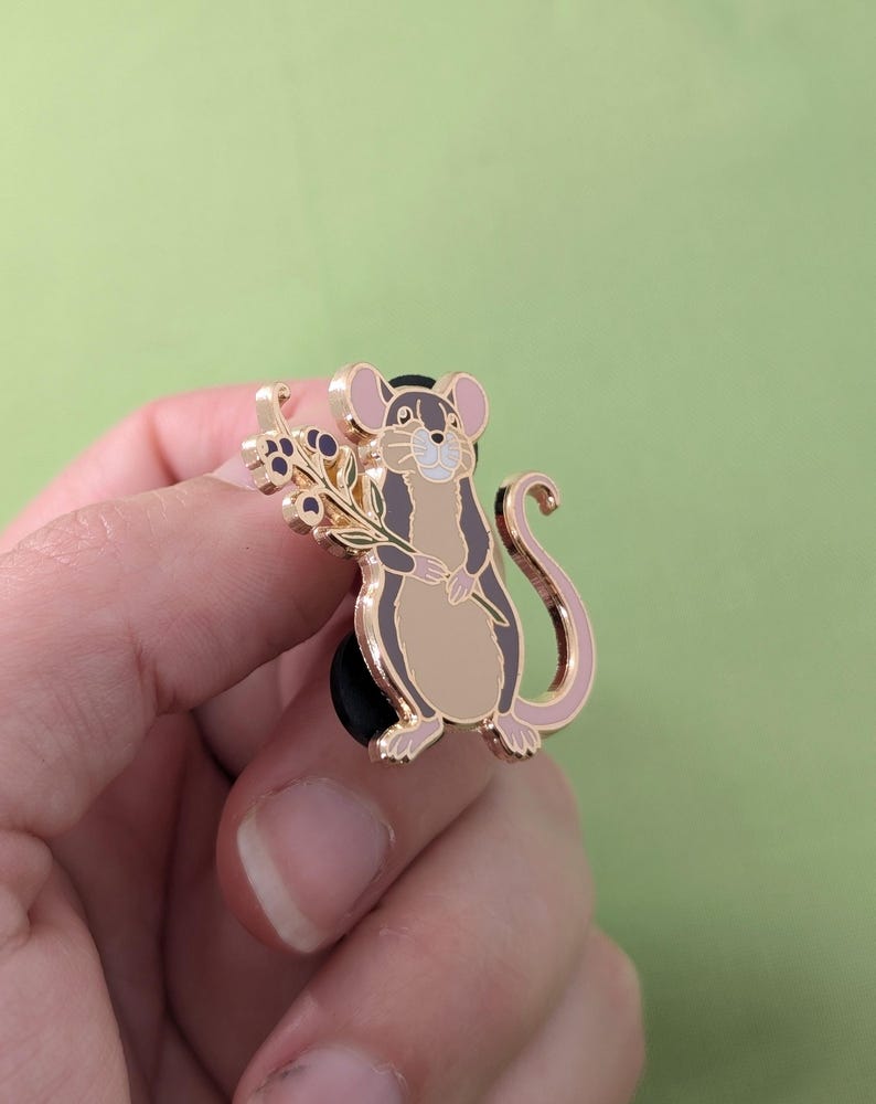 Puede incluir: Pin de esmalte dorado con un rat&oacute;n de dibujos animados sosteniendo una rama de bayas moradas. El rat&oacute;n tiene la nariz y la cola rosas y est&aacute; de pie sobre un respaldo de goma negro.