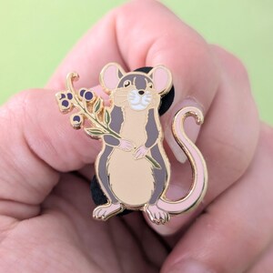Puede incluir: Un pin de esmalte dorado con un rat&oacute;n de dibujos animados sosteniendo una rama de bayas. El rat&oacute;n tiene un cuerpo gris, orejas rosas y una cola rosa.