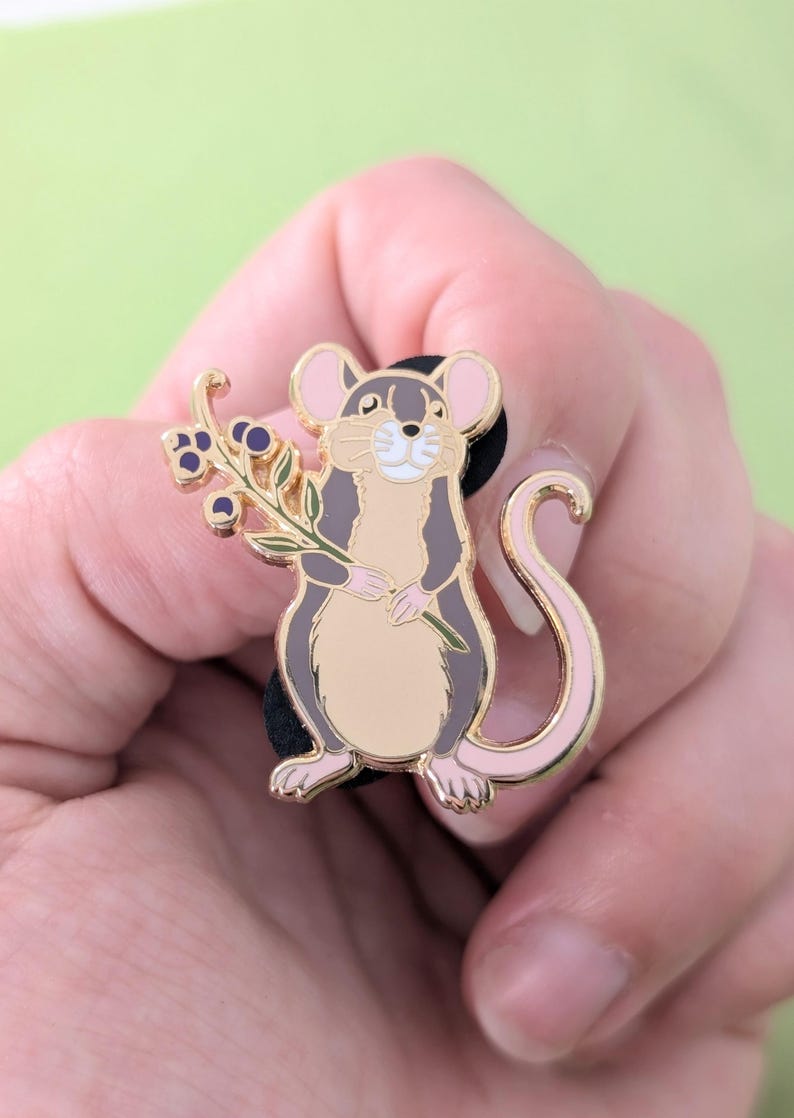 Puede incluir: Un pin de esmalte dorado con un rat&oacute;n de dibujos animados sosteniendo una rama de bayas. El rat&oacute;n tiene un cuerpo gris, orejas rosas y una cola rosa.