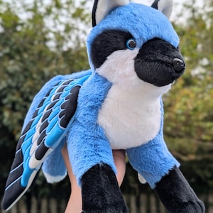 May include: A plush toy in the shape of a blue jay. The stuffed animal has blue, black, and white coloring, with a soft, furry texture. The wings are detailed with black, white, and blue feathers. The toy is held up against a blurred green background.