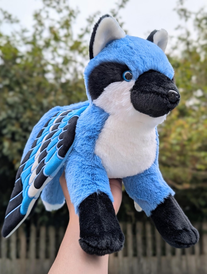 May include: A plush toy in the shape of a blue jay. The stuffed animal has blue, black, and white coloring, with a soft, furry texture. The wings are detailed with black, white, and blue feathers. The toy is held up against a blurred green background.